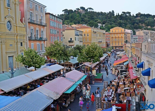 Market happening in Nice. Yellow, Orange and Terracotta houses overlook a street with small trees and market stalls with people shopping