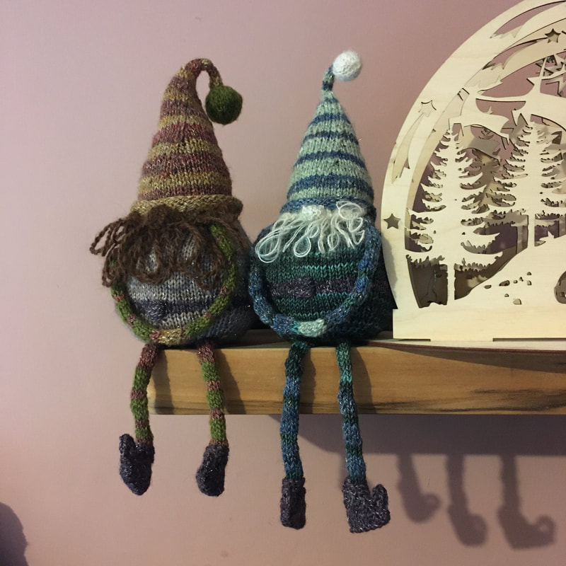 Two striped knitted gnomes sit on a shelf, with their legs dangling. 