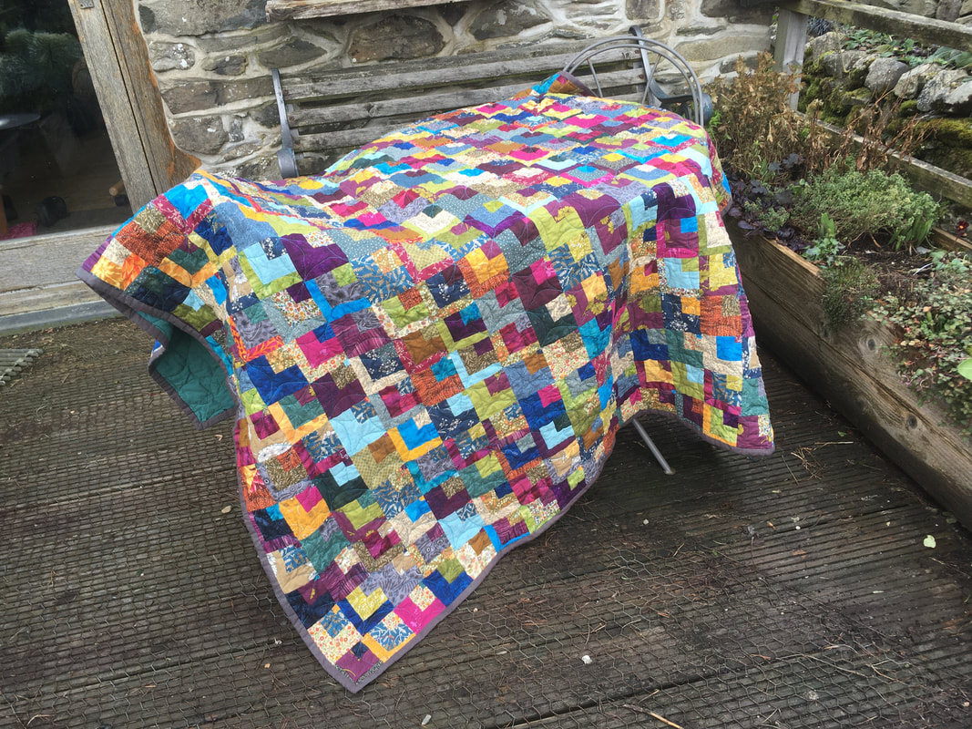A multicoloured quilt draped over a table. The quilt is made from small squares of fabric, and in the top corner of each square is nested a smaller square, and within that an even smaller square. 