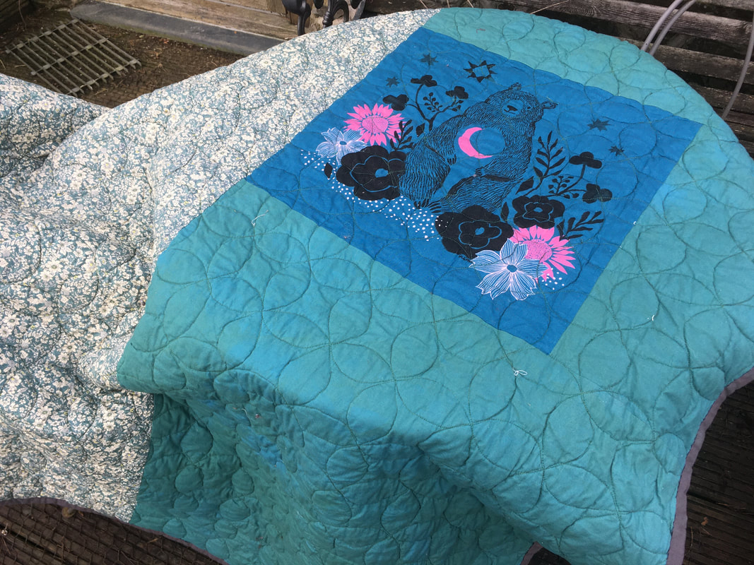 The back of the quilt, half is a white and green floral fabric, the other half plain sea-green, with an inset piece of teal fabric printed with a bear, surrounded by black and pink flowers. 