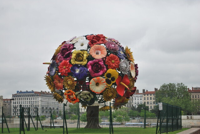 Picture of large metal tree which looks like a ball of flowers, behind a fence on the edge of a river bank