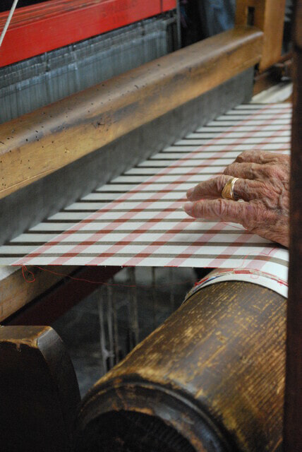 A wrinkled left hand with a wedding ring, resting on weaving on a large loom