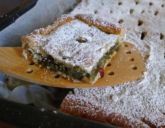 A sort of pie type thing on a wooden cutting board, luted with icing sugar. It kind of looks like pistachio baklava in appearance, there's 2 layers of pastry with something green in the middle