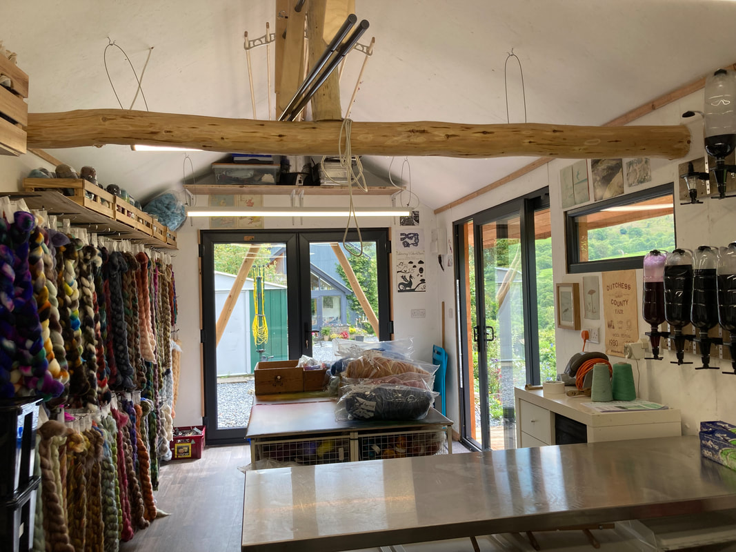 Inside the studio looking out towards the door.  In the foreground a stainless steel table with bar optics containing dye stock solutions on the wall to the right. To the left hang braids of fibre