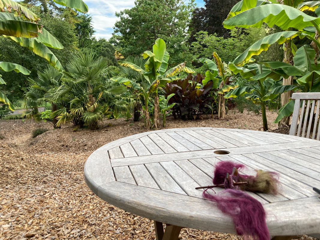 A turkish spindle sits on a wooden table with some purple fibre. In the distances are bananas and palms.