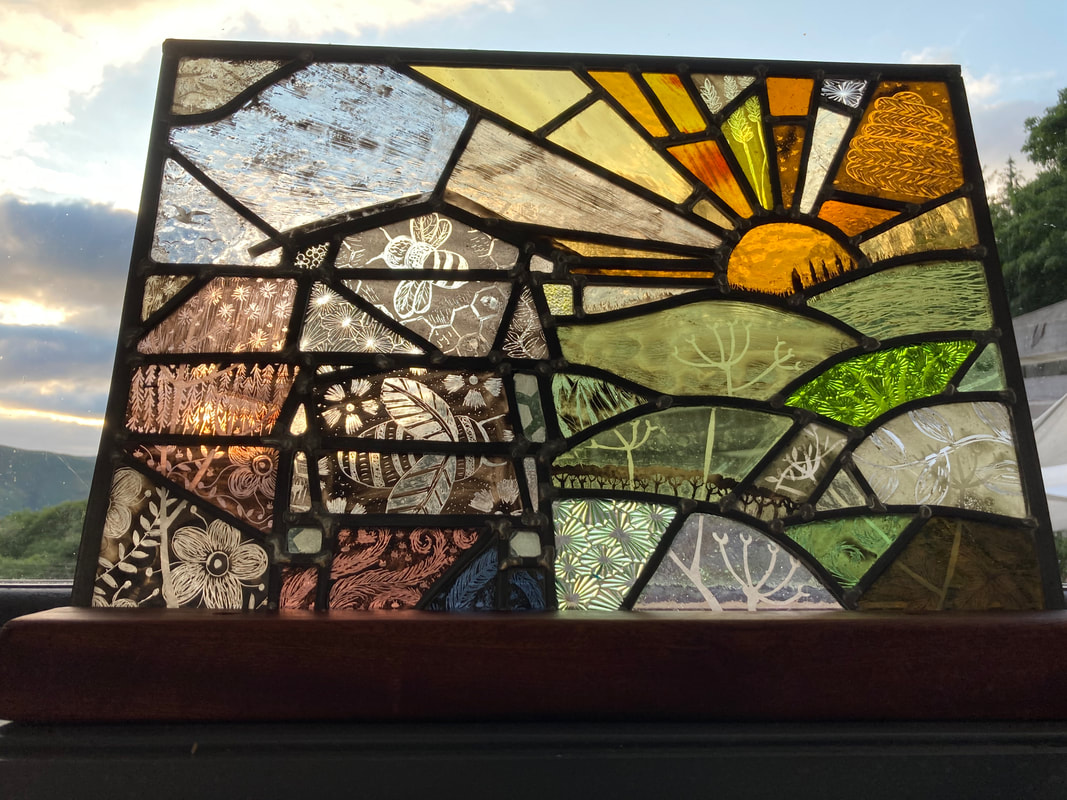 A stained glass panel of a beehive, to the right are green fields made from different shapes and textures of foliage, to the right some pink glass with flower motifs. The beehive has bee motifs on it. At the top is a setting sun. 