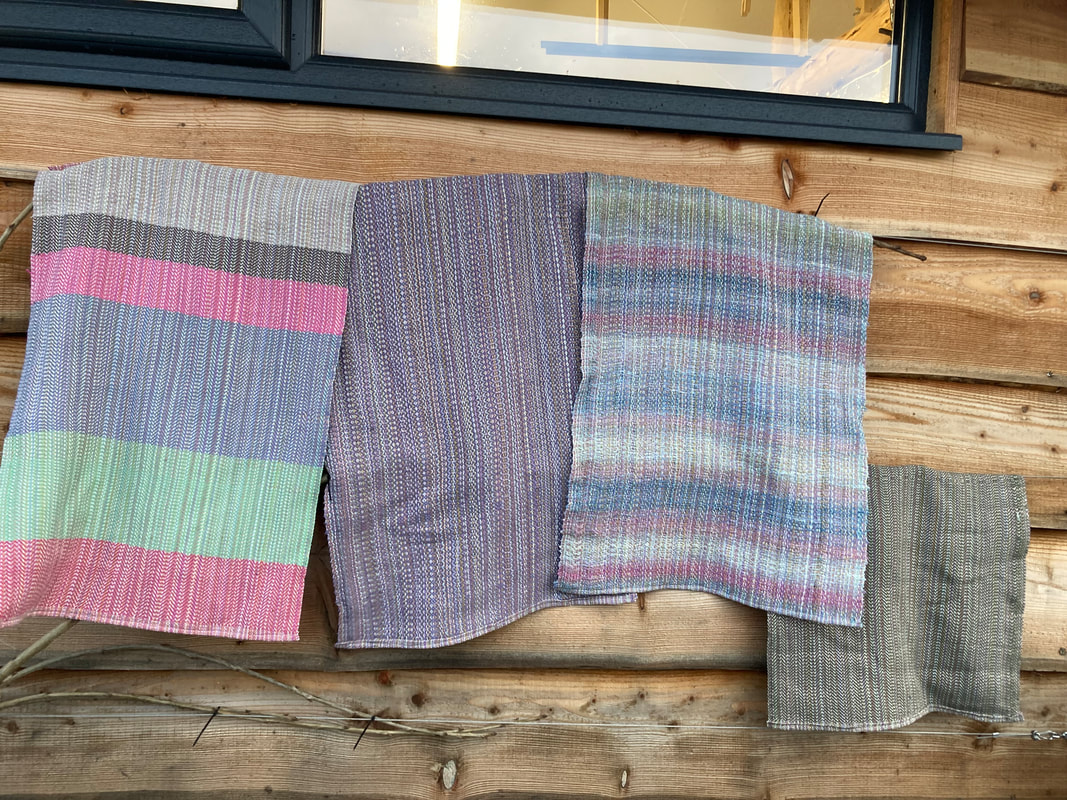 Four tea towels hanging on a line. Left is broad stripes of pink, blue and green, centre uses a mauve weft, thrird is variegated handspun ramie, final is a khaki green weft