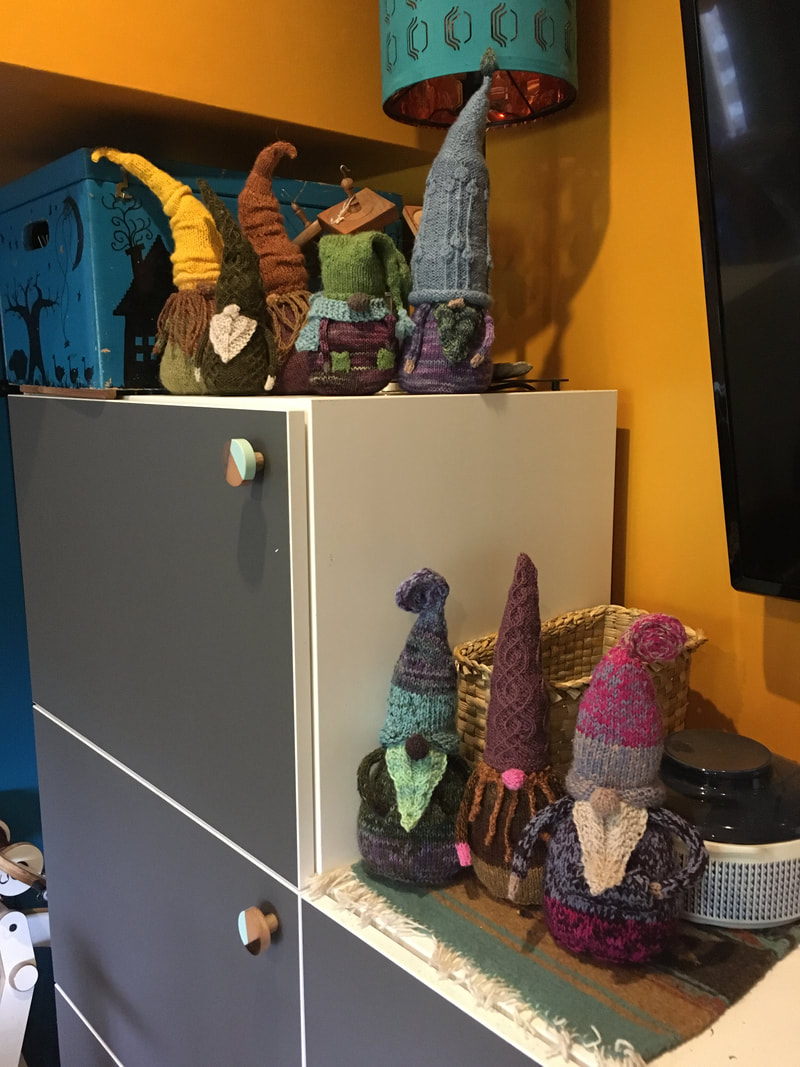 8 knitted gnomes in an assortment of colours sit on 2 shelves. 