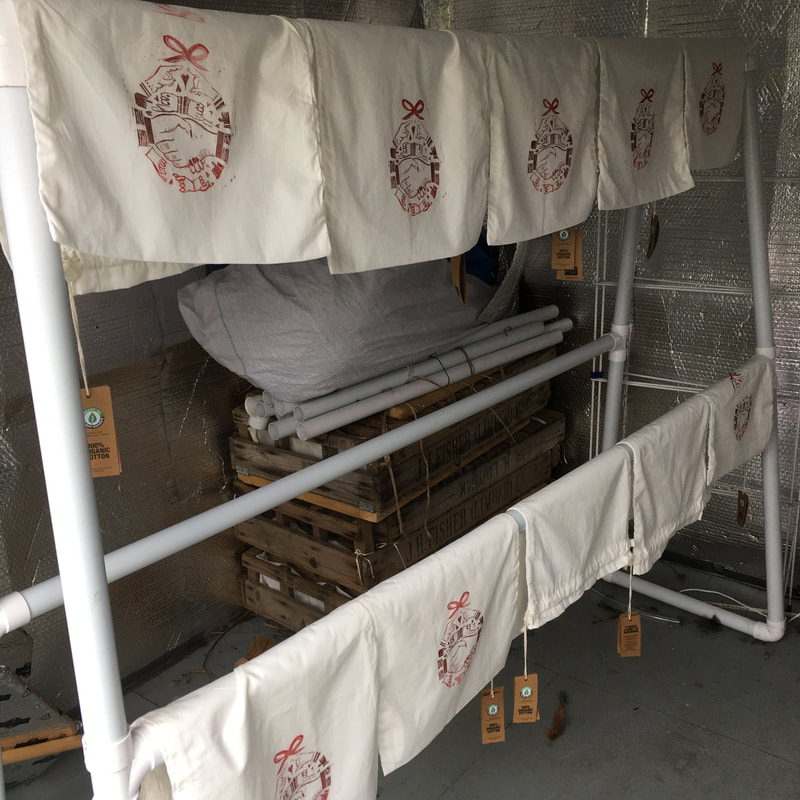 White cotton drawstring bags  hang on a drying rack. They're printed with a red bauble lino cut design  featuring lots of hand outlines