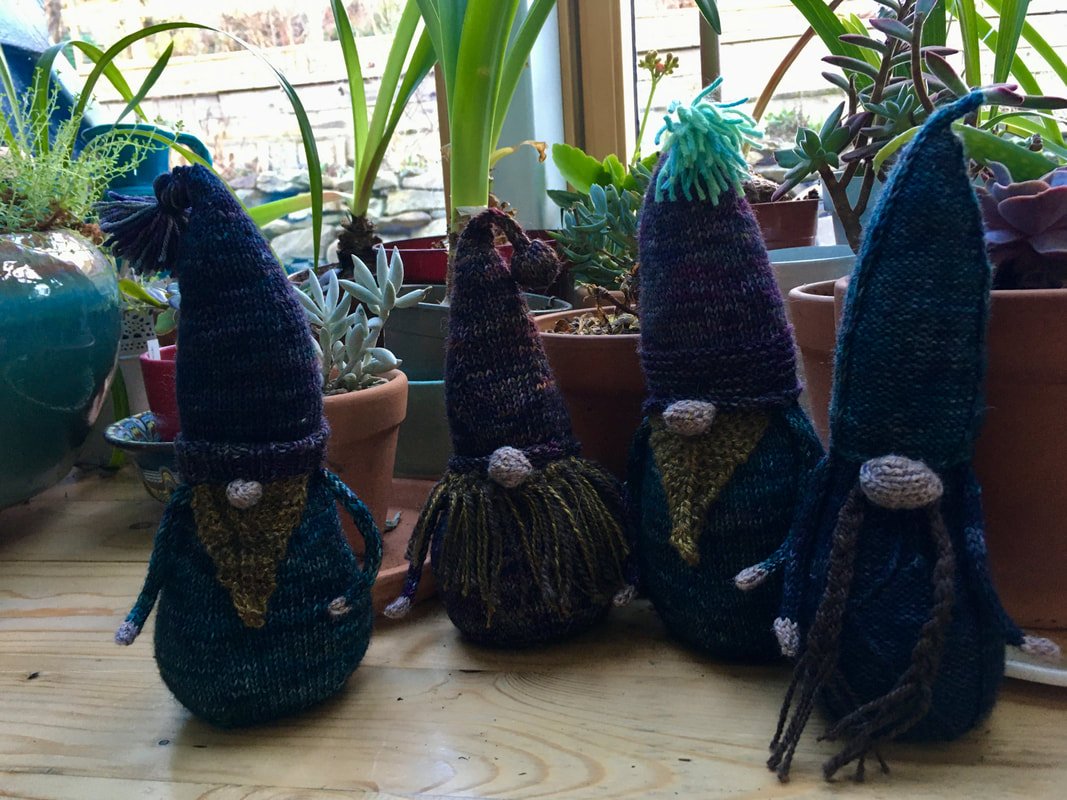 Four knitted gnomes on a wooden windowsill. The gnomes are in dark muted colours with  blonde and blrown beards and round fat caucasian skin toned noses. 